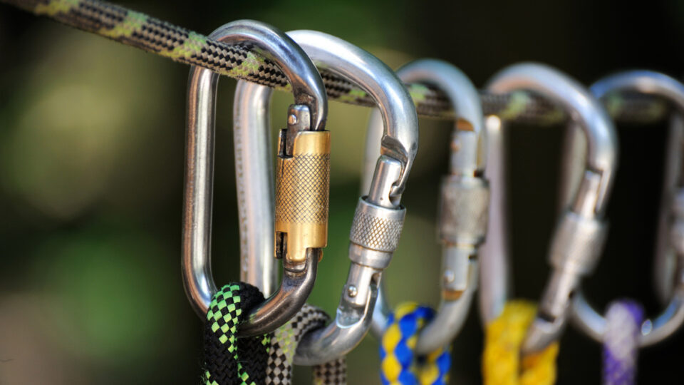 Eine Nahaufnahme von mehreren Karabinern, die an einem Seil hängen. Die Karabiner sind aus Metall und haben verschiedene Designs. Ein Karabiner hat einen goldenen Verschluss, während die anderen silberfarben sind. Die Seile sind in verschiedenen Farben geflochten, darunter schwarz mit grünem Muster, blau und gelb. Der Hintergrund ist unscharf und zeigt eine natürliche Umgebung.