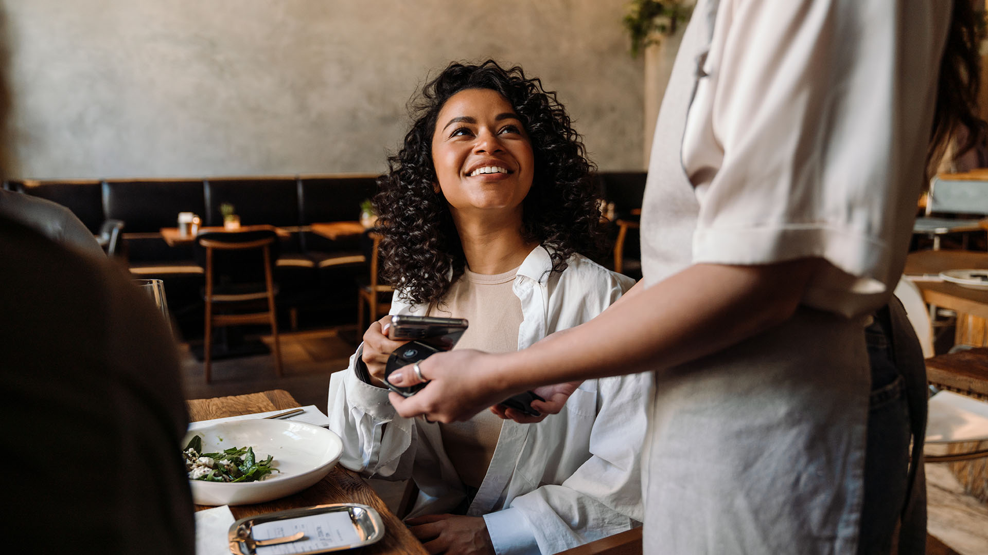 Eine fröhliche Frau mit lockigem Haar sitzt an einem Tisch in einem Restaurant. Sie hält ein Smartphone in der Hand und lächelt, während eine Kellnerin ihr eine Rechnung zeigt. Im Hintergrund sind Tische und Stühle zu sehen.