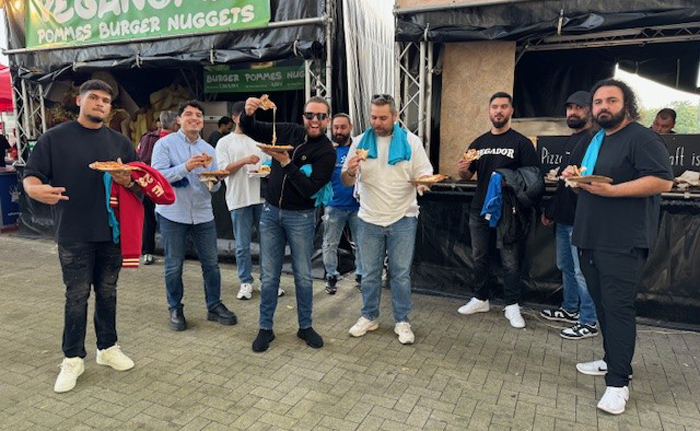 Die Spieler der TARGOBANK Fußballmannschaft essen in dem Fortuna Fußballstadion eine Pizza und schauen dabei in die Kamera.