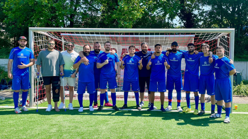 Dreizehn Männer stehen auf einem Fußballfeld vor dem Tor und lächeln in die Kamera.