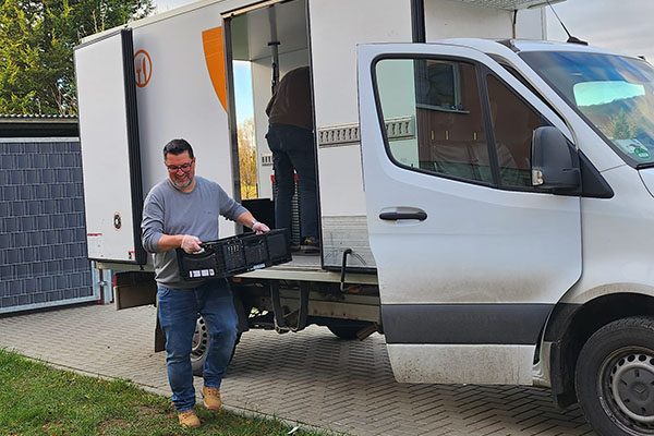 Sascha Watteler trägt eine Kiste der Tafel. Im Hintergrund ist der Transporter der Tafel zu sehen.