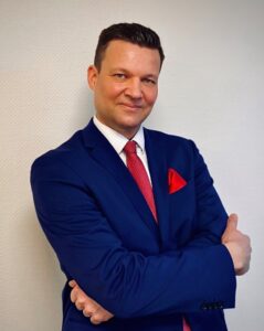 Ein Mann in einem blauen Anzug mit einem roten Krawatte und einem roten Einstecktuch steht mit verschränkten Armen vor einer hellen Wand. Er lächelt und hat kurzes, dunkles Haar.