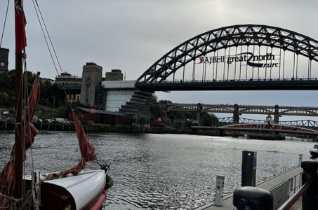 Ein Blick auf den Fluss mit einem Boot im Vordergrund. Im Hintergrund ist die Tyne Bridge mit einer Werbung für den Great North Run zu sehen. Die Uferpromenade ist bebaut, und es sind weitere Brücken sichtbar.