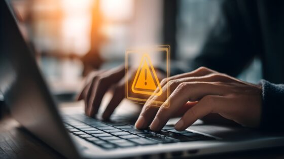 Eine Person tippt auf einer Laptop-Tastatur, während ein gelbes Warnsymbol über dem Bildschirm schwebt. Der Hintergrund ist unscharf und zeigt eine helle, warme Lichtquelle.