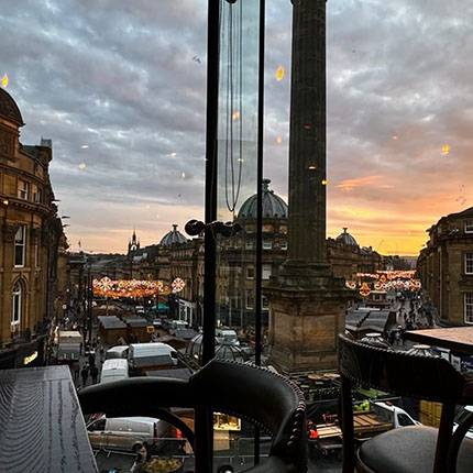 Blick aus einem Fenster auf eine Stadtstraße bei Sonnenuntergang. Die Wolken sind grau und orange, und es sind Lichter zu sehen, die die Straße beleuchten. Autos und Menschen sind sichtbar, und im Hintergrund sind historische Gebäude und eine Säule zu erkennen.