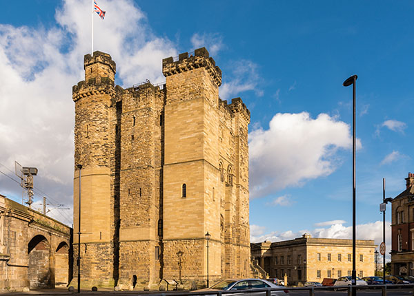 Ein historisches Gebäude, das wie eine Burg aussieht, mit zwei großen Türmen und dicken Mauern. Auf einem Turm weht eine Flagge. Der Himmel ist blau mit einigen Wolken. Im Vordergrund stehen Autos und ein Straßenlaterne ist sichtbar.