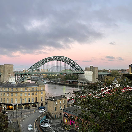 Eine Aussicht auf die Tyne-Brücke in Newcastle, die über den Fluss Tyne führt. Im Vordergrund ist ein historisches Gebäude zu sehen, umgeben von Bäumen und Straßen. Der Himmel ist bewölkt mit sanften Pastellfarben. Links im Bild sind einige Schiffe und die Uferpromenade sichtbar.