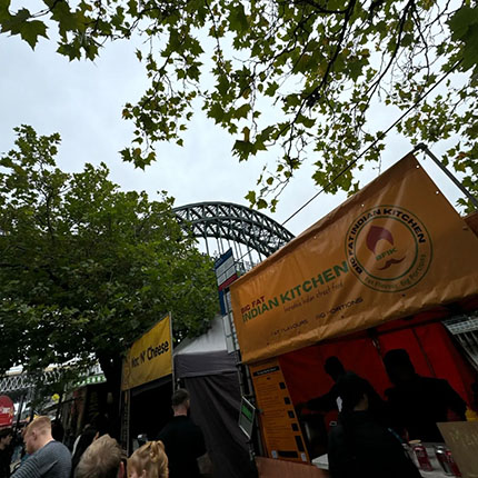 Ein Marktstand mit der Aufschrift 'Indian Kitchen' ist sichtbar, umgeben von Bäumen. Im Hintergrund ist eine große Brücke zu sehen. Der Himmel ist bewölkt.