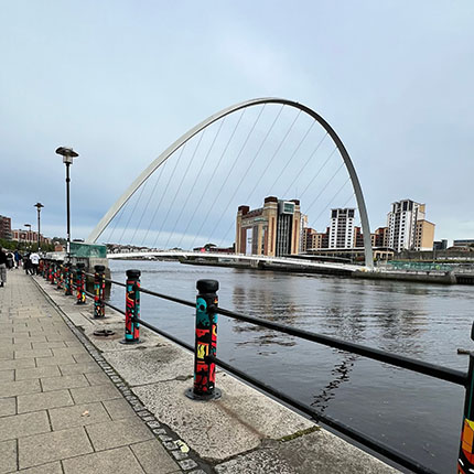 Eine Fußgängerpromenade entlang eines Flusses mit bunten Geländern. Im Hintergrund ist eine große, moderne Brücke mit einem geschwungenen Bogen sichtbar. Am Ufer stehen hohe Gebäude, die das Stadtbild prägen. Der Himmel ist bewölkt und das Wasser des Flusses spiegelt die Umgebung wider.
