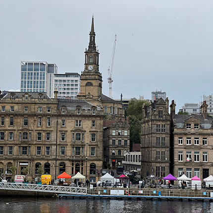 Eine städtische Szene am Wasser mit historischen Gebäuden auf der linken Seite und modernen Hochhäusern im Hintergrund. Es gibt einen markanten Turm mit einer Spitze, und am Ufer sind mehrere Stände mit bunten Markisen zu sehen.
