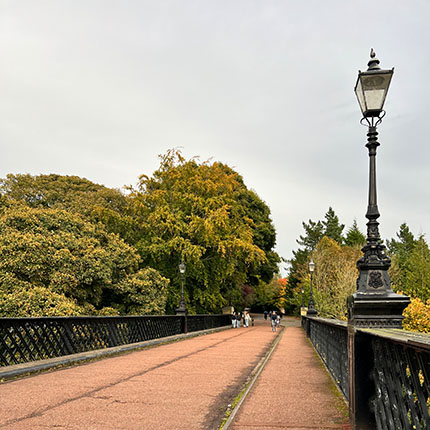 Eine breite Brücke mit einem gepflasterten Weg, umgeben von üppigem Grün und Bäumen, die im Herbstlaub leuchten. Auf der Brücke stehen alte Laternen. Einige Menschen gehen in der Ferne. Der Himmel ist bewölkt.