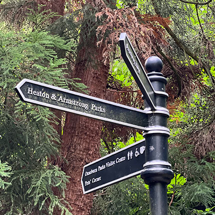 Ein Wegweiser aus schwarzem Metall mit mehreren Pfeilen, die in verschiedene Richtungen zeigen. Die Schilder weisen auf 'Heaton & Armstrong Parks' hin. Darunter steht 'Ouseburn Parks Visitor Centre' und 'Park Café'. Im Hintergrund sind grüne Bäume und Pflanzen zu sehen.
