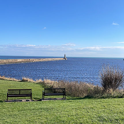Zwei Bänke stehen auf einer grünen Wiese mit Blick auf das Wasser. Im Hintergrund ist ein Leuchtturm an einem Pier sichtbar. Der Himmel ist blau mit einigen Wolken.