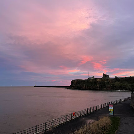 Ein Sonnenuntergang über dem Meer mit sanften rosa und blauen Wolken. Im Hintergrund ist eine Klippe mit einer alten Ruine zu sehen. Ein Leuchtturm steht in der Ferne. Am Ufer verläuft ein Weg mit einem Zaun und einem Schild.