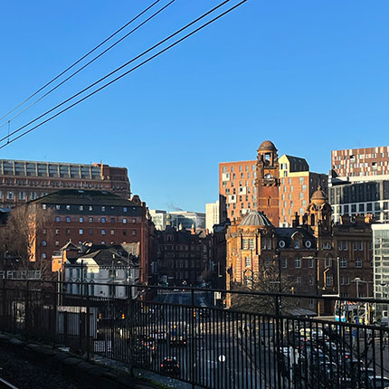 Das Bild zeigt eine städtische Landschaft mit einem klaren blauen Himmel. Im Vordergrund sind Eisenbahnschienen zu sehen, und im Hintergrund ragen verschiedene Gebäude hervor, darunter historische und moderne Architektur. Die Gebäude sind in verschiedenen Farben und Stilen gestaltet, mit einigen, die auffällige Dachstrukturen aufweisen. Die Szene wirkt hell und einladend, mit einer Mischung aus urbanem Leben und architektonischer Vielfalt.