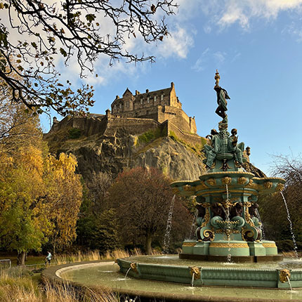 Ein Brunnen im Vordergrund mit einer Statue, umgeben von Bäumen mit herbstlichen Farben. Im Hintergrund sieht man die Burg Edinburgh auf einem Hügel unter blauem Himmel und einigen Wolken.