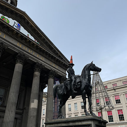 Eine Statue eines Reiters auf einem Pferd, die eine orangefarbene Verkehrscone auf dem Kopf trägt, steht vor einem historischen Gebäude mit Säulen. Der Himmel ist bewölkt.
