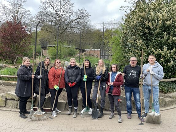 Eine Gruppe von zehn Personen steht im Freien und hält Werkzeuge wie Rechen, Schaufeln und Besen. Sie tragen Handschuhe und sind in legerer Kleidung gekleidet. Im Hintergrund sind Bäume und eine Gartenlandschaft sichtbar.