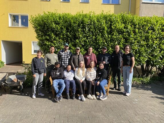 Eine Gruppe von zwölf Personen sitzt und steht vor einem gelben Gebäude. Im Hintergrund ist ein dichter grüner Busch zu sehen. Die Personen tragen legere Kleidung und lächeln in die Kamera. Einige sitzen auf Stühlen, während andere stehen. Im Vordergrund sind zwei Schubkarren sichtbar.