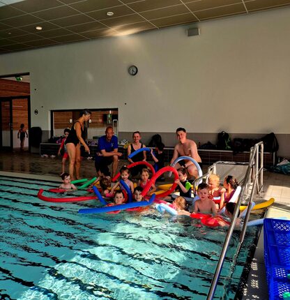 Eine Gruppe von Kindern und Erwachsenen steht am Beckenrand eines Schwimmbads. Die Kinder halten Schwimmnudeln in verschiedenen Farben, während sie im Wasser stehen. Im Hintergrund sieht man eine Trainerin, die mit den Kindern interagiert. Der Bereich ist hell erleuchtet, und eine Uhr ist an der Wand sichtbar.
