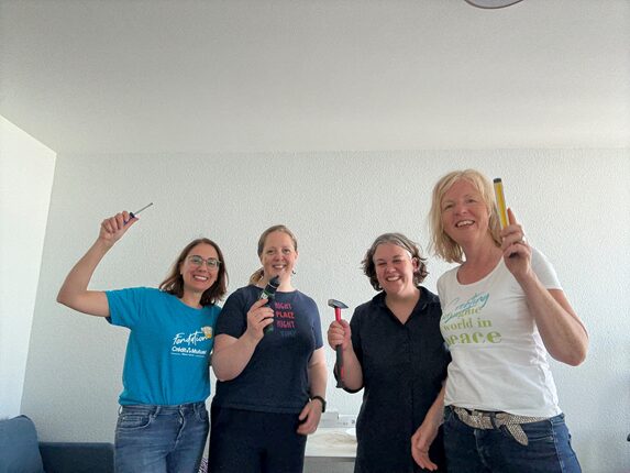Vier Frauen stehen in einem hellen Raum und lächeln. Jede hält ein Werkzeug in der Hand. Die Frau links trägt ein blaues T-Shirt mit dem Text 