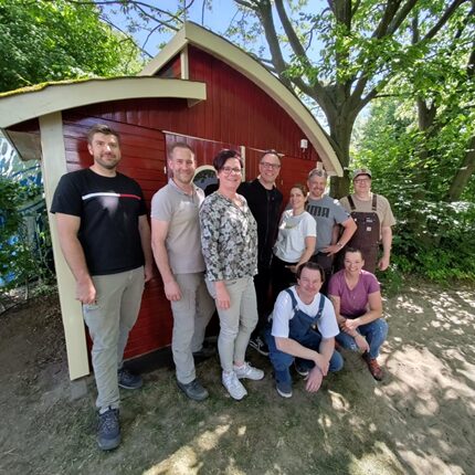 Eine Gruppe von neun Personen steht vor einem roten, hölzernen Gebäude mit einem geschwungenen Dach. Sie lächeln und posieren für das Foto. Im Hintergrund sind Bäume und eine grüne Umgebung sichtbar. Die Personen sind in verschiedenen Kleidungsstilen gekleidet, darunter Hemden und Latzhosen.