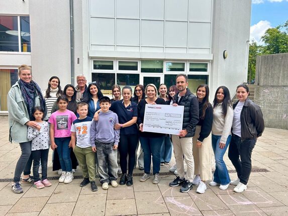 Eine Gruppe von etwa 20 Personen steht vor einem Gebäude und hält einen großen Scheck. Die Gruppe besteht aus Kindern und Erwachsenen, die lächeln. Einige Personen tragen T-Shirts, andere sind in Freizeitkleidung. Im Hintergrund sind große Fenster und eine graue Wand sichtbar. Der Scheck ist auf den Namen 