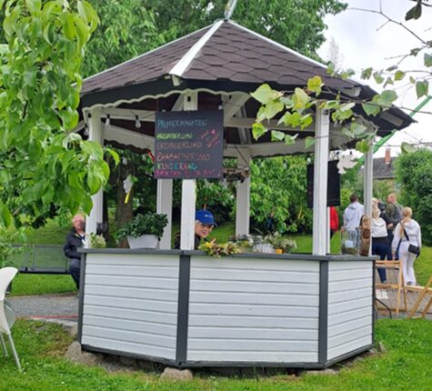 Ein Pavillon mit einem Holzstand, der grüne Pflanzen und eine Tafel mit Angeboten zeigt. Im Hintergrund sind Menschen zu sehen, die sich unterhalten. Es ist bewölkt.