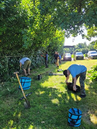 Eine Gruppe von Menschen arbeitet im Freien, umgeben von hohen Bäumen. Einige Personen beugen sich über den Boden, während andere mit Werkzeugen beschäftigt sind. Auf dem Gras stehen mehrere blaue Eimer, und im Hintergrund sind Autos zu sehen. Es ist ein sonniger Tag mit viel grünem Laub.