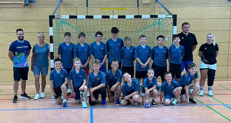 Eine Gruppe von 20 Jungen und Mädchen, die in blauen Sporttrikots gekleidet sind, posiert in einer Sporthalle. Sie stehen vor einem Handballtor und lächeln in die Kamera. Einige von ihnen sitzen, andere stehen, während Trainer und Betreuer am Rand stehen. Die Umgebung zeigt einen Holzboden und eine Wand mit einem Netz, das das Handballtor umgibt.