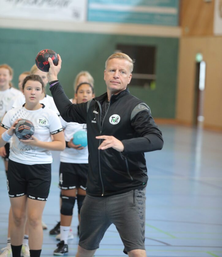Ein Trainer in einem Sportanzug steht in einer Sporthalle und wirft einen Handball. Hinter ihm stehen mehrere Mädchen in weißen Trikots, die ebenfalls Handbälle halten. Die Halle hat einen Holzboden und grüne Wände.
