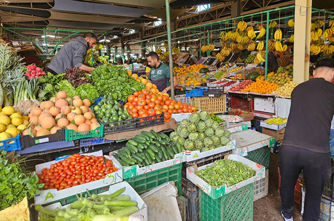 Ein belebter Markt mit verschiedenen Obst- und Gemüsesorten. Verkäufer sortieren frisches Gemüse wie Gurken, Tomaten und grüne Blätter. Im Hintergrund hängen Bananen und weitere Früchte sind sichtbar. Die Waren sind in Kisten und Körben angeordnet, und mehrere Kunden sind ebenfalls unterwegs.