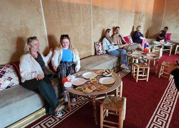 Eine Gruppe von Menschen sitzt in einem gemütlichen Raum mit braunen Wänden und roten Teppichen. Zwei Frauen in bequemen Outfits lachen und sitzen auf einem Sofa. Auf dem Tisch vor ihnen sind verschiedene traditionelle Speisen angerichtet, darunter kleine Teller mit Dips und Brot. Im Hintergrund sitzen weitere Personen, die in entspannter Atmosphäre essen und plaudern.