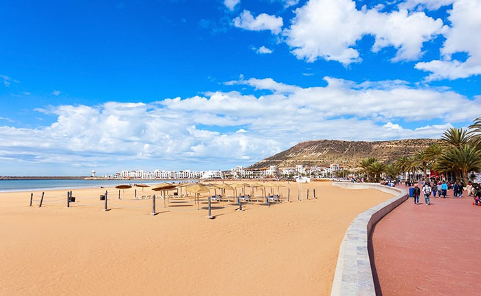 Ein sonniger Strand mit feinem Sand erstreckt sich unter einem blauen Himmel voller Wolken. Im Hintergrund sind Gebäude und Palmen zu sehen. Am Strand stehen einige Sonnenschirme und Liegen, während eine Promenade mit Spaziergängern verläuft.