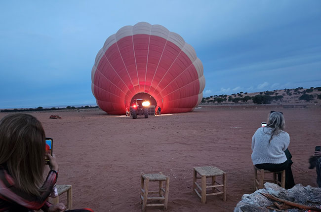 Ein Heißluftballon wird auf einem sandigen Boden aufgeblasen, während einige Personen auf Stühlen sitzen und Fotos machen. Der Himmel ist bewölkt und es wird langsam dunkel.