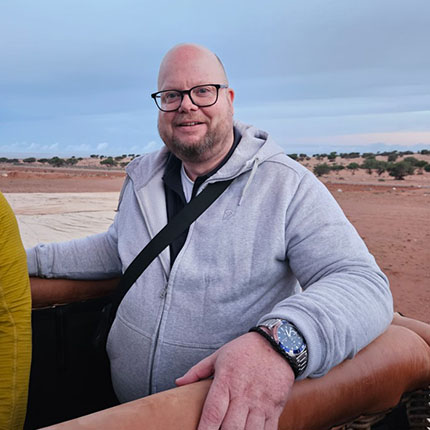 Ein Mann mit Brille und Glatze steht in einem Heißluftballonkorb. Er trägt einen grauen Hoodie und hat eine schwarze Umhängetasche. Im Hintergrund ist eine wüstenartige Landschaft mit einigen Bäumen und einem bewölkten Himmel zu sehen.