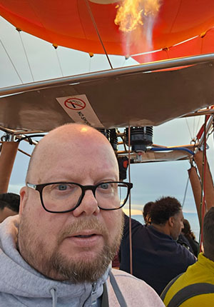 Ein Mann mit Glatze und Brille steht in einem Heißluftballon. Im Hintergrund sieht man andere Passagiere und die Flamme, die aus dem Brenner des Ballons kommt. Der Himmel ist bewölkt.