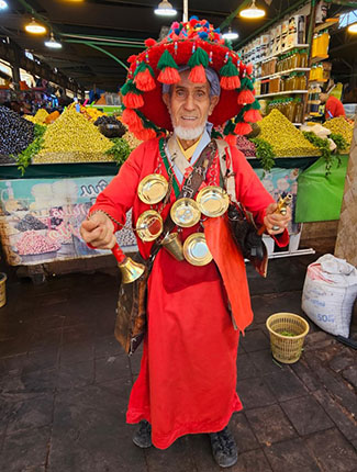 Ein älterer Mann trägt ein auffälliges rotes Gewand und einen bunten Hut mit Quasten. Er steht in einem Markt mit bunten Waren im Hintergrund. Um seinen Hals hängen mehrere kleine Kannen und Glocken. Der Mann lächelt und zeigt seine Hände, während er in die Kamera blickt.
