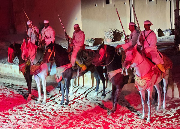 Fünf Reiter in traditioneller Kleidung sitzen auf ihren Pferden. Die Reiter tragen weiße Hemden und Kopfbedeckungen. Die Atmosphäre ist durch rotes Licht beleuchtet. Die Pferde sind geschmückt, und der Boden ist mit Sand bedeckt.
