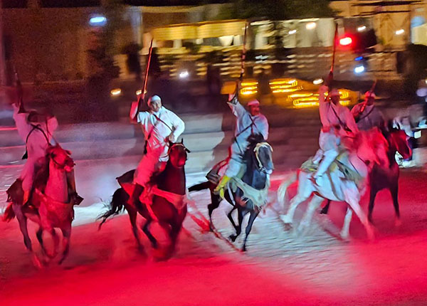 Sechs Reiter in traditioneller Kleidung reiten auf Pferden. Sie tragen Lanzen und sind von rotem Licht umgeben, was eine dynamische Atmosphäre schafft. Im Hintergrund sind unscharfe Lichter von Gebäuden zu sehen.
