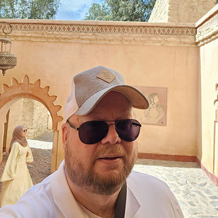 Ein Mann mit einer Baseballkappe und Sonnenbrille steht im Vordergrund vor einer hellen Wand mit einem Bogen. Im Hintergrund ist eine Frau in einem langen, beigen Kleid sichtbar, die durch einen architektonischen Durchgang geht. Die Umgebung ist sonnig und zeigt eine traditionelle, dekorative Architektur.