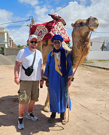 Ein Mann steht neben einem Kamel, während ein weiterer Mann in traditioneller Kleidung daneben steht. Der Mann trägt ein blaues Gewand und eine Kopfbedeckung, während der andere in einem weißen T-Shirt und Shorts gekleidet ist. Das Kamel hat eine dekorative Decke auf dem Rücken. Im Hintergrund sind einige Gebäude und ein blauer Himmel mit Wolken zu sehen.