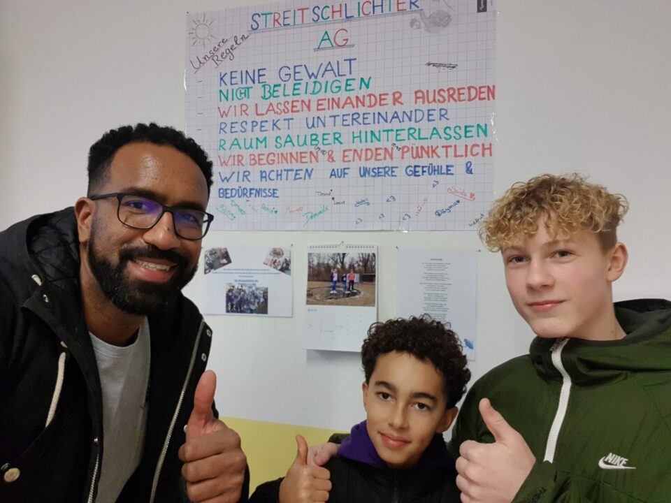 Drei Personen posieren lächelnd vor einem Poster mit Regeln der Streitschlichter AG. Der Mann links trägt eine Brille und hat einen Bart, während die beiden Jungen, einer mit lockigen Haaren und der andere mit glattem Haar, Daumen hoch zeigen. Das Poster enthält Regeln wie 