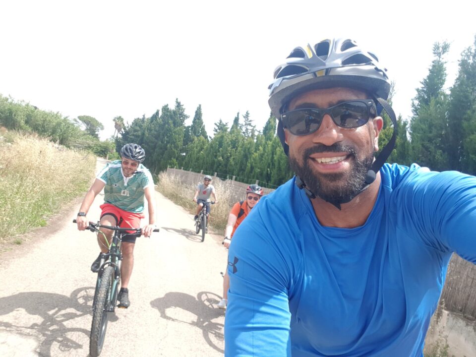 Eine Gruppe von Radfahrern auf einem schmalen, staubigen Weg. Im Vordergrund lächelt ein Mann mit dunkler Hautfarbe, Brille und Fahrradhelm in die Kamera. Er trägt ein blaues T-Shirt. Hinter ihm fahren drei weitere Radfahrer: ein Mann in einem gestreiften Hemd und roten Shorts, sowie zwei weitere Männer in sportlicher Kleidung. Die Umgebung ist grün mit Bäumen und hohem Gras.