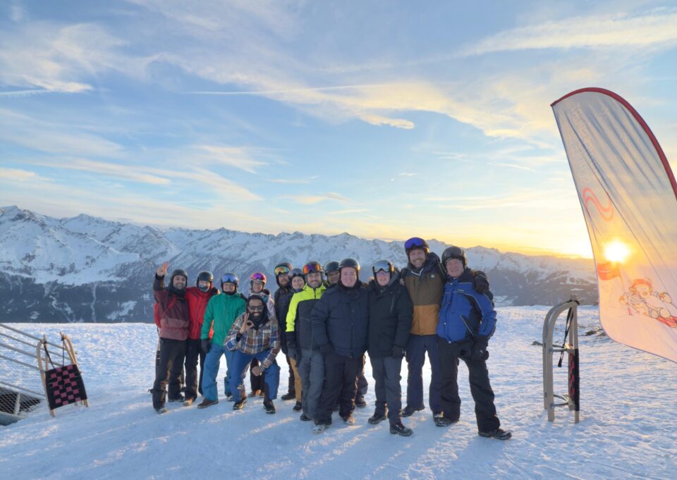 Eine Gruppe von 14 Personen steht auf einem schneebedeckten Berg mit Blick auf schneebedeckte Berge im Hintergrund. Einige tragen Skiausrüstung und Helme. Die Sonne geht hinter den Bergen unter und der Himmel ist blau mit Wolken. Auf der rechten Seite ist ein Banner mit einem Bild eines Schneemanns und dem Text 