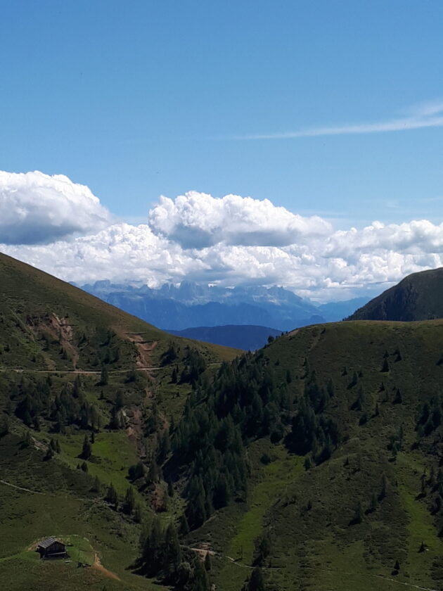 Eine malerische Berglandschaft mit sanften Hügeln, grünen Wiesen und verstreuten Bäumen. Im Hintergrund sind hohe Berge und eine klare blaue Himmel mit weißen Wolken zu sehen. Ein kleines Holzhaus steht in der unteren linken Ecke der Szene.