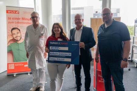 Eine Gruppe von vier Personen steht zusammen, während eine Frau einen großen Scheck von 10.000 Euro präsentiert. Der Scheck ist für die Westdeutsche Spenderzentrale/DRK. Im Hintergrund ist ein Werbeplakat von WSZE zu sehen. Die Personen tragen unterschiedliche Kleidung: ein Arzt in weißem Kittel, eine Frau in rotem Shirt, und zwei weitere Männer in Geschäfts- und Freizeitkleidung.