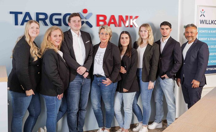 Eine Gruppe von acht Personen steht in einem modernen Büro der TARGOBANK. Die Personen tragen schwarze Blazer und Jeans. Die Frau in der Mitte hat blonde Haare und trägt ein T-Shirt mit der Aufschrift 