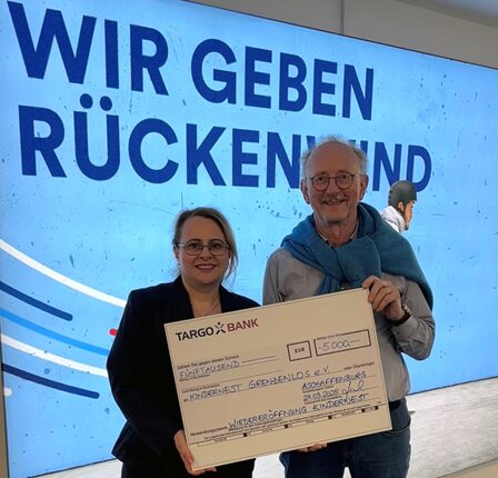 Eine Frau und ein älterer Mann halten einen großen Scheck vor einer Wand mit dem Text 