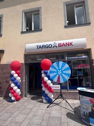 Eine TARGO BANK-Filiale mit einem großen Eingang. Vor dem Eingang stehen zwei Säulen aus roten, weißen und blauen Ballons. Neben den Ballons steht ein Glücksrad mit blauer und weißer Farbgestaltung. Die Fenster sind hell und freundlich.